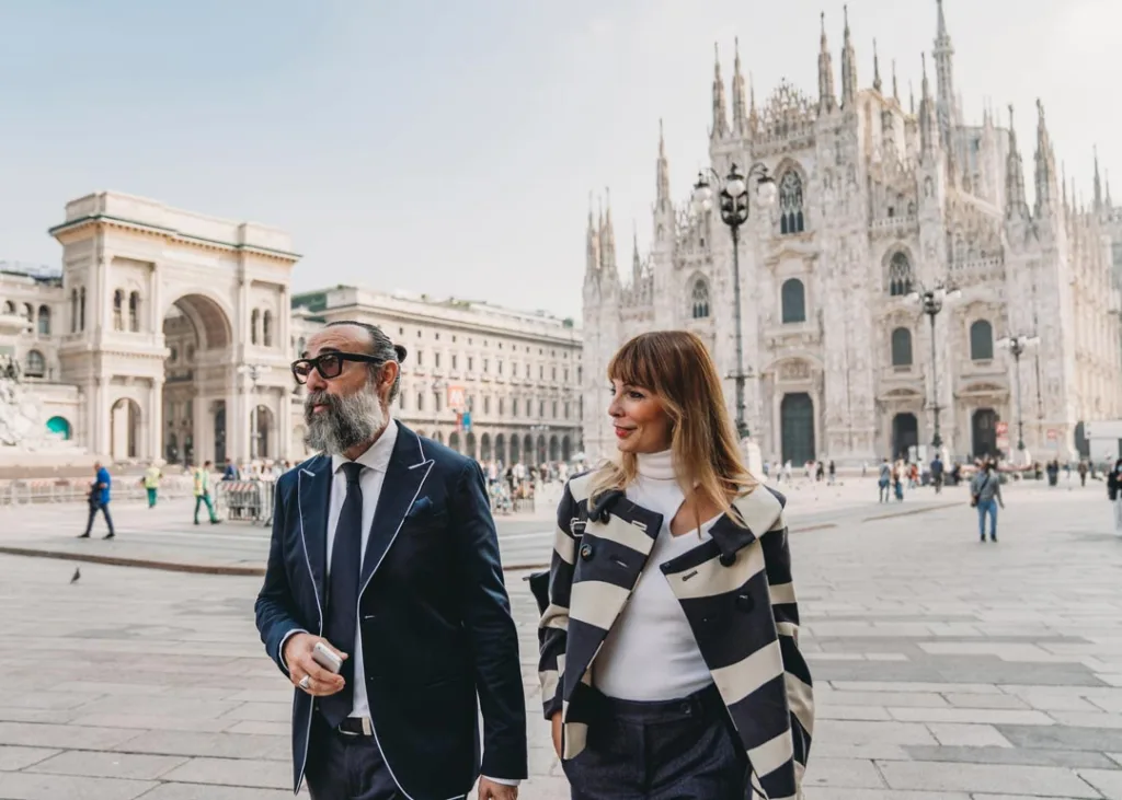 Un uomo con barba e codino cammina con una donna su una piazza italiana.